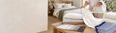 Textile specialist inspecting white bed linen beside folded sheets, towels and fabric samples in a bright Australian-style bedroom.
